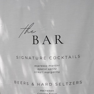 Może przedstawiać: Biały lniany szyld barowy z napisem "the BAR" w eleganckim czarnym skrypcie. Poniżej wymienione są charakterystyczne koktajle, takie jak espresso martini i aperol spritz, a także piwa, twarde seltzery i wina.