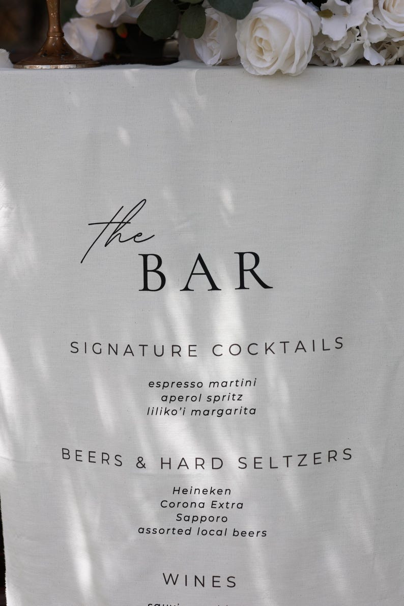 Może przedstawiać: Biały lniany szyld barowy z napisem "the BAR" w eleganckim czarnym skrypcie. Poniżej wymienione są charakterystyczne koktajle, takie jak espresso martini i aperol spritz, a także piwa, twarde seltzery i wina.