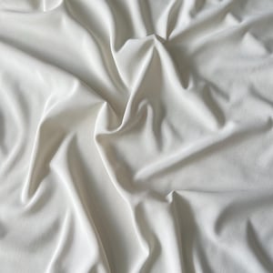 May include: A close-up shot of a white fabric with soft folds and shadows. The fabric appears to be smooth and flowing, with a subtle sheen. The overall impression is one of elegance and simplicity, suitable for apparel or home decor.
