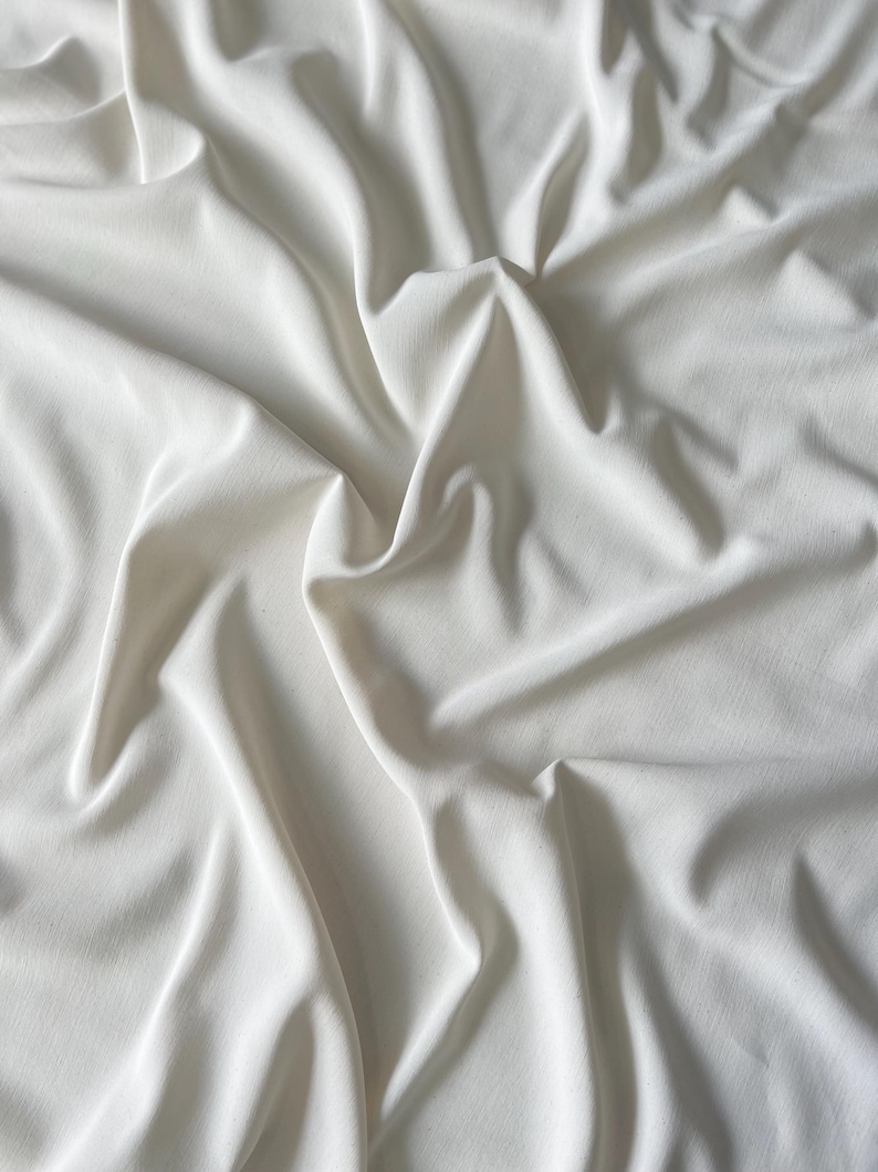 May include: A close-up shot of a white fabric with soft folds and shadows. The fabric appears to be smooth and flowing, with a subtle sheen. The overall impression is one of elegance and simplicity, suitable for apparel or home decor.