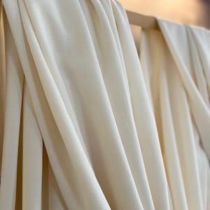 May include: Cream-colored fabric hanging from a wooden rail. The material is soft and drapes in flowing folds, creating a play of light and shadow. The fabric's neutral tone suggests it could be used for clothing or interior design purposes.