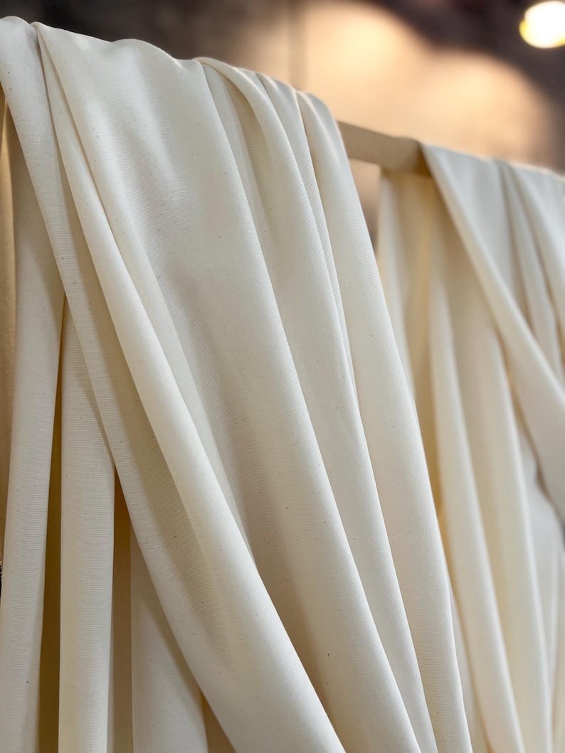 May include: Off-white fabric draped over a wooden rod. The fabric has a soft texture and falls in gentle folds, creating shadows and highlights. The neutral color suggests a natural material, possibly linen or cotton, suitable for clothing or home decor.