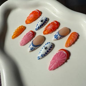May include: A collection of artificial nails in various designs. Some nails are orange, resembling citrus fruit slices, while others are pink with a floral pattern. Several nails feature a white base with blue floral designs, and some have a nude base with blue accents.