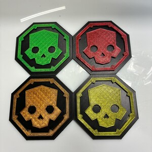 May include: Four black coasters with a skull design. Each coaster has a different colored skull: green, red, gold, and yellow. The skull is inside a black octagon.