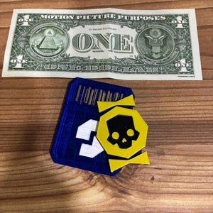 May include: A blue and white SD card with a yellow skull and crossbones design, next to a one-dollar bill. The dollar bill has the text "MOTION PICTURE PURPOSES" and "ONE DOLLAR". The objects are on a wooden surface.