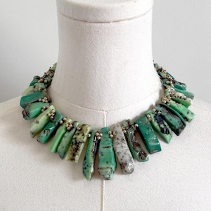 May include: A statement necklace featuring a series of raw, cylindrical green stone segments with dark inclusions, interspersed with small metallic beads. The stones vary in shades of mint, forest green, and white, arranged in a dense, collar-style design.