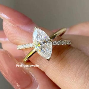 Puede incluir: Anillo de compromiso con diamante talla marquesa y banda dorada. El anillo presenta un gran diamante talla marquesa transparente como pieza central, con diamantes más pequeños engastados en la banda. El anillo está sobre un fondo neutro.
