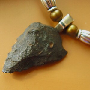 Può includere: Una collana con un ciondolo a forma di punta di freccia in pietra grigio scuro dalla consistenza ruvida, sospeso a una serie di perline color argento metallizzato e perline rotonde e lisce color bronzo. La punta di freccia è appuntita.