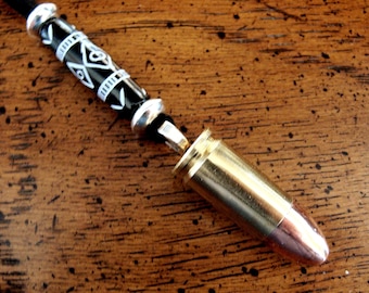 Può includere: Un ciondolo per collana composto da un bossolo metallico con punta color rame, fissato a un cordino nero decorato con una perla cilindrica nera con motivi geometrici bianchi e distanziatori in metallo argentato su ciascun lato.