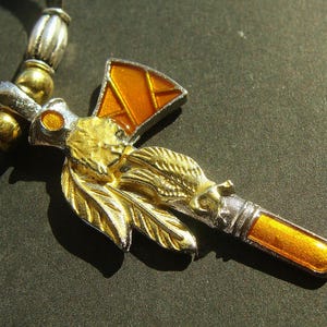 May include: A gold and silver-toned pendant necklace with an orange enamel accent. The pendant features a Native American profile, feathers, and a tomahawk design. The necklace has a black cord and silver and gold beads.
