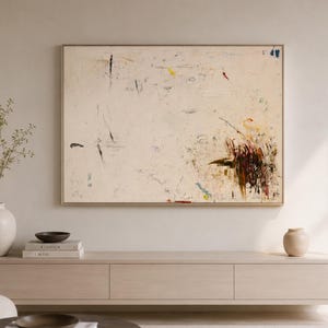 May include: Abstract painting with a cream-colored background and a gold frame. The artwork features brushstrokes in black, red, blue, and yellow. The painting hangs above a light wood cabinet with decorative items.