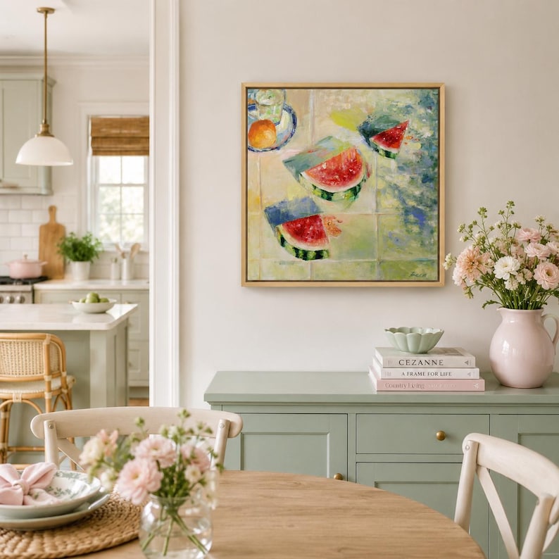 May include: A square framed painting depicting slices of watermelon and a small orange fruit on a light-coloured surface. The artwork features vibrant red, green, and yellow brushstrokes, displayed on a wall above a light green wooden cabinet.