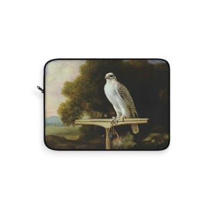 May include: Laptop sleeve featuring a detailed painting of a falcon perched on a wooden stand. The bird has white and gray plumage, set against a backdrop of trees and a landscape. The sleeve has a black zipper and a black border.
