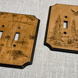 May include: Two rectangular wooden light switch plates with dark edges. The left plate features two bird illustrations in a field. The right plate displays a forest scene with three pine trees and a bird flying above, both with two switch cutouts.