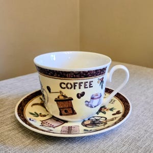 May include: A cream-colored ceramic teacup and saucer set featuring a brown border of coffee beans. The cup displays the word COFFEE in black letters, accompanied by illustrations of a vintage coffee grinder, a mug, and a coffee press.