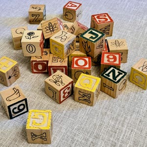 May include: A collection of wooden alphabet and number blocks featuring various printed illustrations including a bee, a flower, a bird, and scissors. Each cube displays letters, numbers, and symbols in black, red, green, and yellow ink on natural wood.