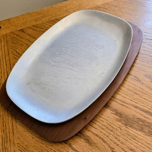 May include: A silver-colored, oval-shaped serving tray sits atop a dark brown wooden tray. The silver tray has visible scratches. The wooden tray has rounded corners and a smooth surface. The trays are on a wooden table.