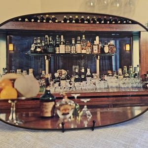May include: An oval serving tray with a dark wood frame and light-colored woven handles on each side. The surface features a printed image of a bar interior with shelves of various glass bottles, glassware, and a bowl of fruit on a wooden counter.