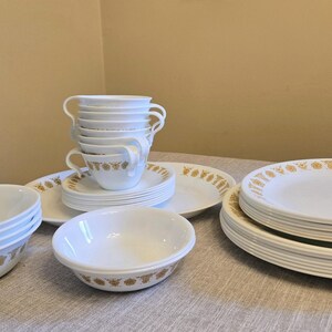 May include: A collection of white dinnerware featuring a repeating gold floral pattern along the rims. The set includes stacked dinner plates, salad plates, bowls, and teacups with handles, all displaying a consistent gold botanical motif.