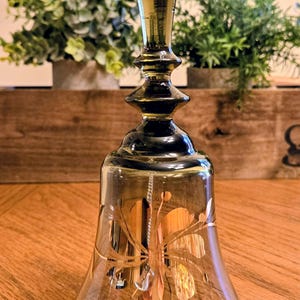 May include: A decorative glass bell with a dark amber hue and a gold-colored floral design. The bell has a long, ornate handle and a clear clapper. The bell is sitting on a wooden surface.