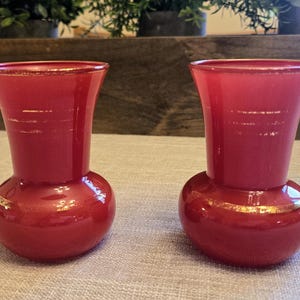 May include: Two red glass vases with a bulbous base and flared top. The vases are a vibrant red color and have a smooth, glossy finish. They are likely decorative items, possibly for flowers or display.