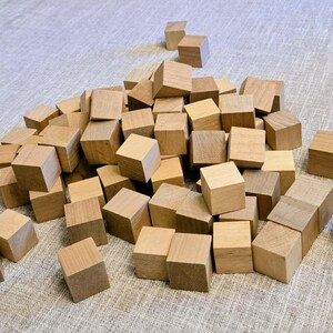 May include: A large collection of small, light brown wooden cubes scattered in a pile. Each cube features a smooth, unfinished surface with visible natural wood grain patterns. The blocks are uniform in shape and size, creating a textured arrangement.