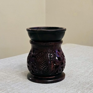 May include: A dark brown stone oil burner featuring a smooth, circular top bowl resting on a base with intricate, hand-carved paisley and floral cutouts. The structure is cylindrical with a flared bottom rim, showcasing a polished finish.