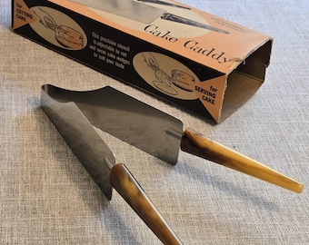 May include: A stainless steel cake caddy utensil with two hinged blades and brown marbled handles, presented with its original cardboard box labeled Hostess Cake Caddy, featuring illustrations for cutting and serving cake wedges.