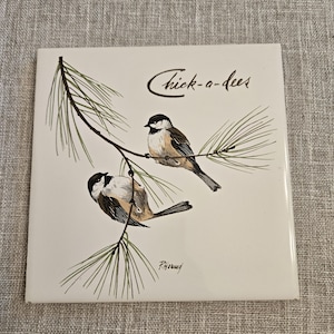 May include: A square white ceramic tile featuring a detailed illustration of two black-capped chickadees perched on a green pine branch. The text Chick-a-dees is printed in a dark script font above the birds, with the artist signature P. Horned below.