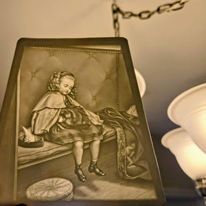May include: A lampshade featuring a sepia-toned illustration of a child with curly hair sitting on a tufted bench, wearing a cape and holding a small animal. The scene includes a round woven object on the floor and a draped fabric beside the child.