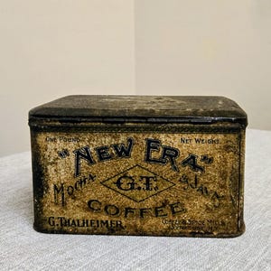 May include: A rectangular vintage metal coffee tin with a distressed gold and black finish. The front features the text "New Era" Coffee, "G.T.", "Mocha & Java", "One Pound Net Weight", and "G. Thalheimer Coffee & Spice Mills, Syracuse, N.Y."