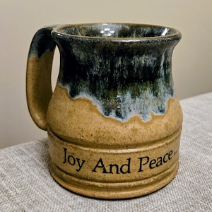 May include: A handmade ceramic mug with a large handle. The mug features a two-tone design, with a tan base and a dark green and blue glaze at the top. The words "Joy And Peace" are printed in black on the mug's body. The mug is sitting on a neutral-colored surface.