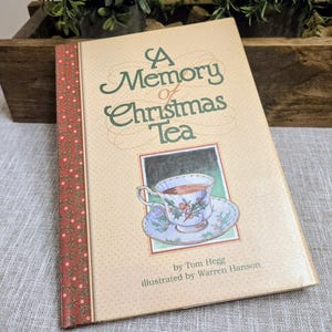May include: A Memory of Christmas Tea book with a beige cover and a decorative border. The cover features the title in green script, an illustration of a teacup, and the author and illustrator's names.