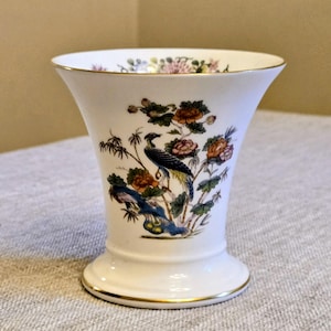 May include: A white porcelain vase with a flared rim and a circular base, featuring a gold-trimmed edge. The exterior is decorated with a colorful illustration of a bird perched among flowering branches and foliage in shades of blue, orange, and green.