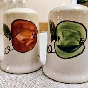 May include: A pair of ceramic salt and pepper shakers. One shaker features a red apple design, the other a green apple. Both have a beige base color with black outlines and green leaves. The apple designs are hand-painted.