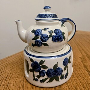 May include: A white ceramic teapot with a matching warmer. The teapot and warmer are decorated with blue floral designs and green leaves. The teapot has a lid and a handle. The warmer has cut-out sides.