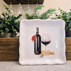 May include: White square ceramic plate with a wavy edge, featuring a wine bottle, a glass of red wine, corks, and a wax seal design. The plate is set against a backdrop of greenery and wine glasses.
