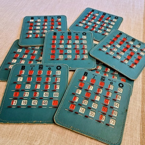 May include: A collection of seven teal bingo cards featuring a grid of numbers with sliding red plastic shutters. Each card displays the word BINGO at the top, numbered squares, and the text MADE IN U.S.A. printed near the top edge.