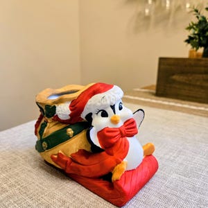 May include: A ceramic Christmas decoration featuring a penguin wearing a Santa hat and a red bow tie, sitting on a red sled. The penguin is next to a tan sack with green and gold accents.