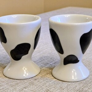 May include: A pair of white ceramic egg cups featuring a black cow-print pattern. Each cup has a flared base and a smooth, glossy finish, designed to hold a single egg upright. The cups are identical in shape and size, showcasing a simple, functional design.