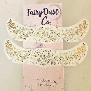 May include: Four white patches with gold star and snake designs are displayed on a pink box. The box has the text "Fairy Dust Co." and "*Includes 2 Patches". The patches are decorated with gold stars and a gold snake.