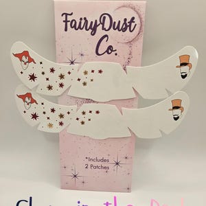 May include: Two white under-eye patches featuring a clown graphic on the left and a top-hatted figure on the right, with scattered star designs. The packaging is labelled Fairy Dust Co. and states Includes 2 Patches and Glow in the Dark.
