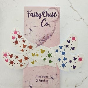 May include: A package of Fairy Dust Co. patches with a pink background and the text "Fairy Dust Co." in purple script. The patches are white with colorful Mickey Mouse-shaped paint splatters. The package includes two patches.