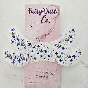 May include: A pink box with the text "Fairy Dust Co." and star graphics. White patches with blue star and green circle designs are arranged around the box. The text "*Includes 2 Patches" is also visible.