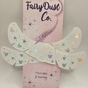Könnte beinhalten: Eine rosa Verpackung mit Fairy Dust Co. Patches, mit dem Text "Fairy Dust Co." in Lila. Die weißen Patches haben schillernde Mickey Mouse-Kopfformen und Sterne. Die Verpackung enthält "Includes 2 Patches".