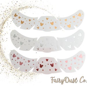 Könnte beinhalten: Drei Sätze weißer, dekorativer, augenförmiger Aufkleber. Jeder Satz hat eine andere Metallic-Farbe: Gold, Silber und Roségold. Die Aufkleber sind mit Mickey Mouse-Silhouetten und Sternen verziert. Das FairyDust Co. Logo befindet sich unten rechts.
