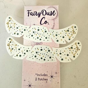 May include: A package of two white eye patches with a star and bow and arrow design, with the text "Fairy Dust Co." in a cursive font. The patches have a wing-like shape and are decorated with green and gold stars.