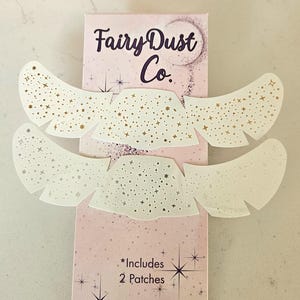 May include: Two white, star-studded wing-shaped patches with gold accents are displayed over a pink box. The box reads "Fairy Dust Co." and "*Includes 2 Patches."