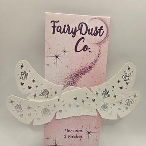 以下が含まれることがあります： 「Fairy Dust Co.」の文字と星のアクセントが入ったピンク色の箱。城、木、地球儀のデザインが施された白いパッチが箱の前に表示されています。「*Includes 2 Patches」の文字も表示されています。