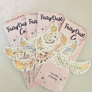 Peut inclure: Plusieurs paquets roses de patchs "Fairy Dust Co." avec des motifs d'étoiles et de paillettes. Les paquets indiquent "Fairy Dust Co." et "*Includes 2 Patches". Les patchs ont des détails dorés, argentés et à paillettes.